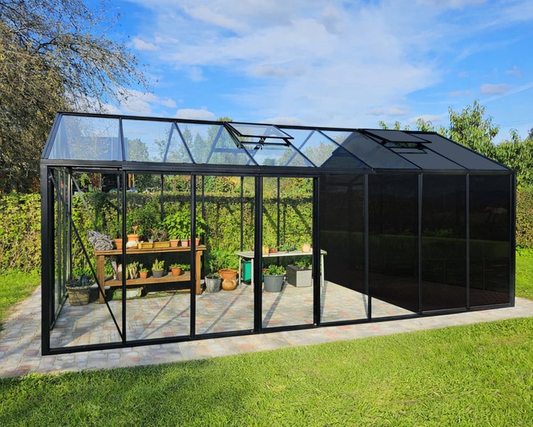 Großes Gewächshaus mit schwarzem Metallrahmen und Glasscheiben, stehend in einem Garten auf einer gepflasterten Fläche.