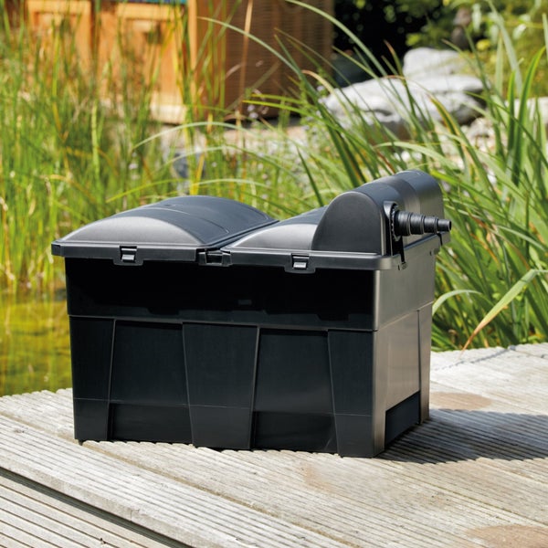 Teichfilter aus schwarzem Kunststoff auf einem Holzdeck neben einem bepflanzten Gartenteich.