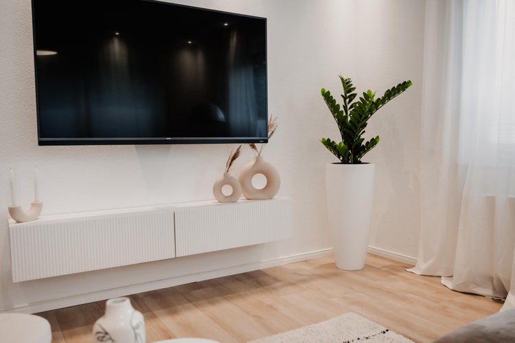Modernes helles Wohnzimmer mit flachem Fernseher an weißer Wand, weißem schwebendem Lowboard, hohem Pflanztopf mit Grünpflanze und Holzboden.