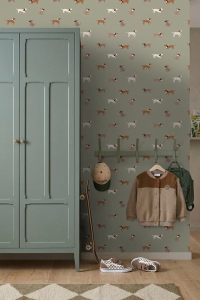 Vliestapete mit Hundemuster im Kinderzimmer neben einem grünen Schrank, einer Garderobe mit Jacke und einem Skateboard auf Holzfußboden.