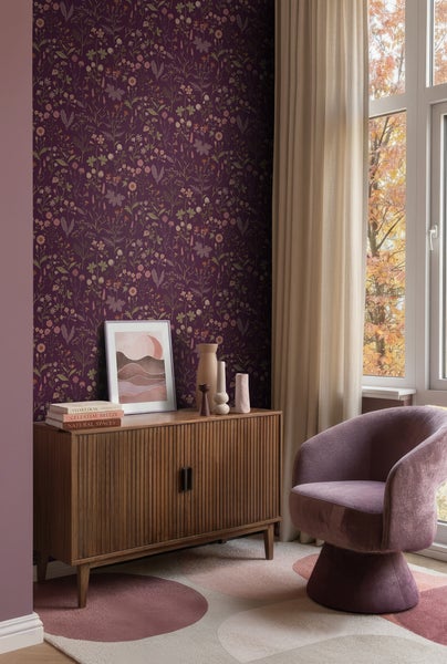 Zimmer mit dunkelvioletter Blumentapete, Sideboard aus Holz, lila Samtsessel und hellen Vorhängen an einem Fenster mit Herbstbäumen.