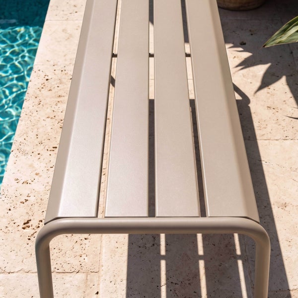 Moderne Gartenbank aus Metall mit Längslatten neben einem Swimmingpool auf einer Steinterrasse.