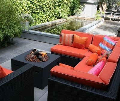 Moderner Garten-Loungebereich mit Ecksofa aus schwarzem Geflecht, orangen Polstern und quadratischer Feuerstelle auf einer Steinterrasse am Teich.