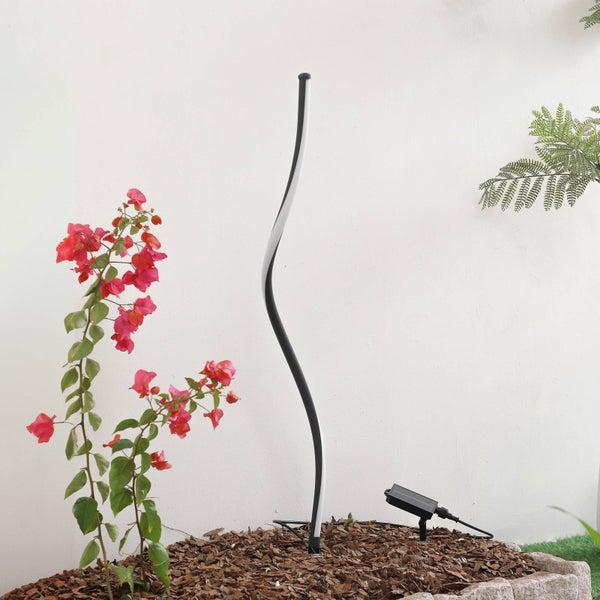 Geschwungene LED-Gartenleuchte in einem Beet mit Rindenmulch und rosa Blüten vor einer hellen Wand.