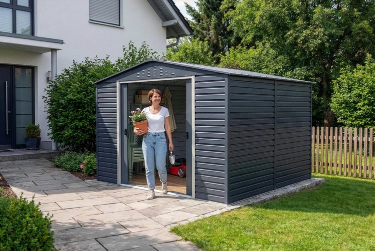 Anthrazitfarbenes Metall-Gerätehaus mit Schiebetür und Satteldach in einem hellen Garten neben einem Wohnhaus mit Steinweg.