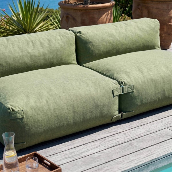 Grünes modulares Outdoor-Lounge-Sofa aus strukturiertem Stoff auf einer Holzterrasse mit Pflanzen und Wasser im Hintergrund.