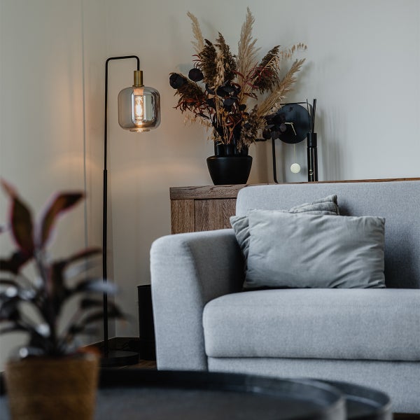 Modernes Wohnzimmer mit grauem Sofa, einer Stehlampe mit Glasschirm und Trockenblumen in einer schwarzen Vase auf einer Holzkommode.
