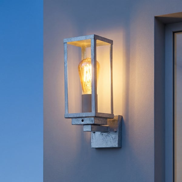 Rechteckige Außenwandleuchte aus verzinktem Stahl mit Glaseinsätzen und leuchtender Filamentlampe an einer Wand.
