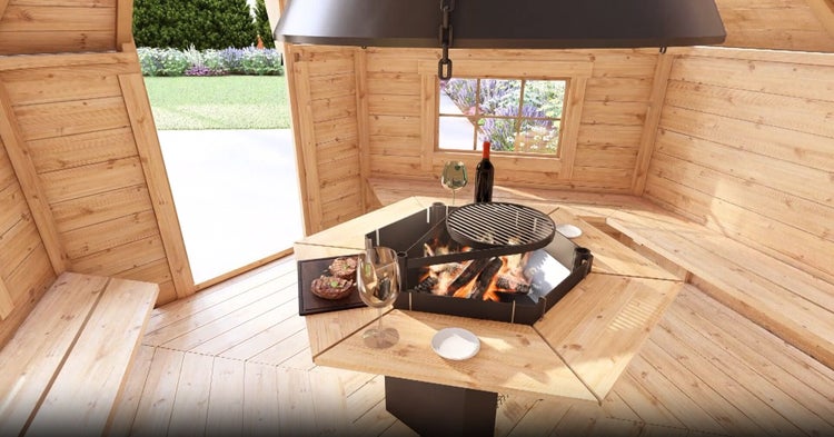Innenansicht einer Grillkota aus Holz mit zentraler Feuerstelle, Grillrost, umlaufendem Holztisch, Sitzbänken und Abzugshaube.