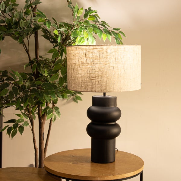 Dekorative Tischleuchte mit beigem Lampenschirm und schwarzem Lampenfuß auf einem runden Tisch.