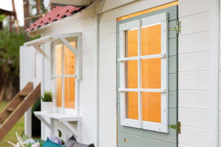 Weißes Kinderspielhaus aus Holz mit grauer Tür, Sprossenfenster und warmer Innenbeleuchtung im Garten.