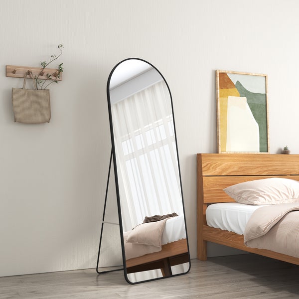 Großer Standspiegel mit bogenförmigem Abschluss und schmalem schwarzem Rahmen in einem hellen Schlafzimmer neben einem Holzbett.