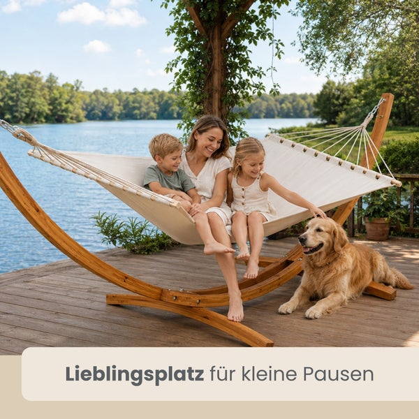 Frau und zwei Kinder sitzen in einer beigen Hängematte mit Holzgestell auf einem Steg am See, daneben liegt ein Hund.