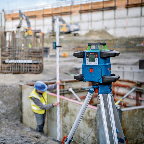 Rotationslaser auf Stativ auf einer Baustelle mit Bauarbeiter und Bagger im Hintergrund