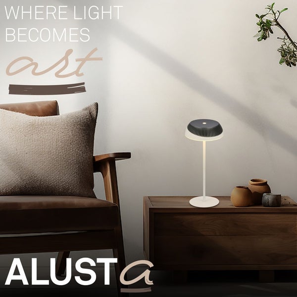 Moderne Alusta Tischleuchte auf einer Holzkommode neben einem braunen Sessel in einem hellen Wohnraum.