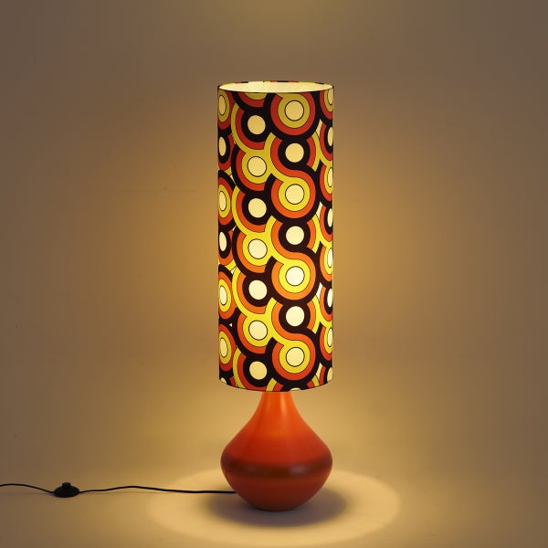 Retro-Tischleuchte mit zylinderförmigem Schirm im 70er-Jahre-Muster und bauchigem, orangefarbenem Fuß, eingeschaltet.