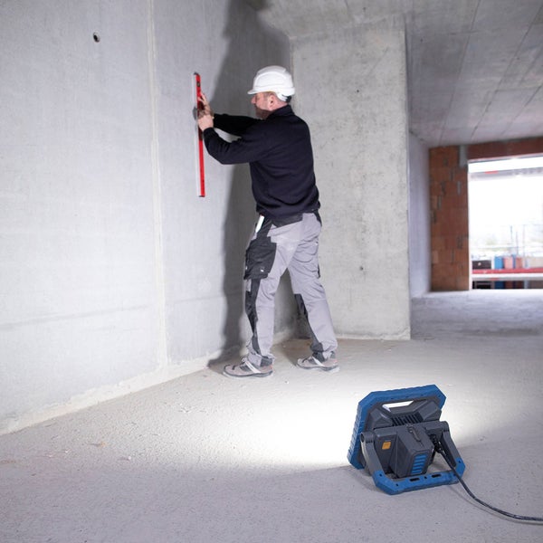 Ein Bauarbeiter mit Wasserwaage an einer Wand in einem Rohbau, im Vordergrund eine Baustellenleuchte.