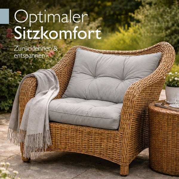 Geflochtener Gartensessel mit grauen Polstern und einer Decke in einem hellen Garten mit passendem Beistelltisch.