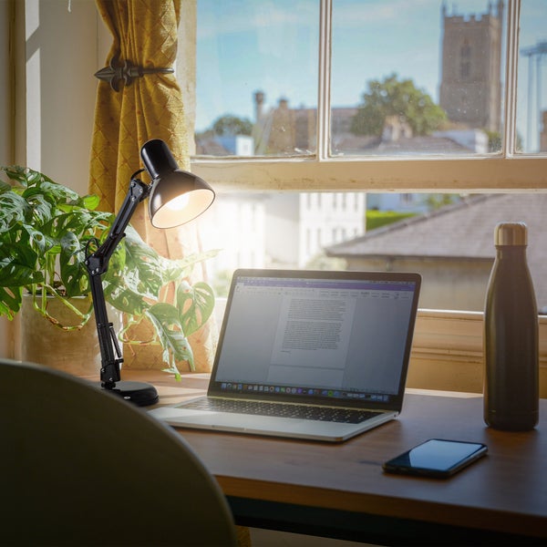 Schwarze Schreibtischleuchte mit Gelenkarm auf einem Holztisch neben einem Laptop und einer Zimmerpflanze vor einem hellen Fenster.
