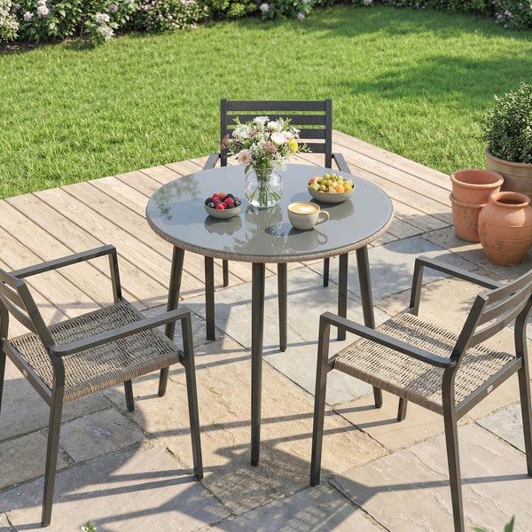 Modernes Gartenmöbel-Set mit rundem Glastisch und drei Stühlen auf einer Terrasse aus Holz und Stein neben einer gepflegten Rasenfläche.