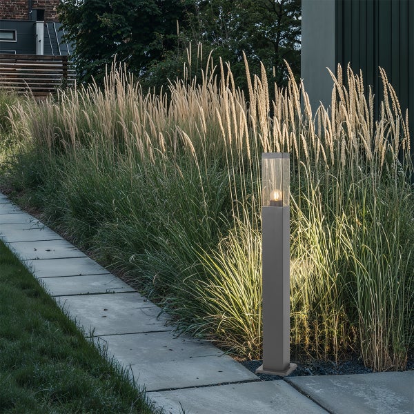 Moderne Außen-Sockelleuchte aus Metall und Glas beleuchtet einen Steinweg im Garten neben hohen Ziergräsern.