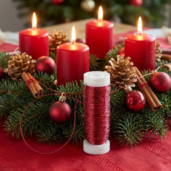 Eine Spule mit feinem Bindedraht vor einem Adventskranz mit vier brennenden Kerzen, Dekokugeln, Zimtstangen und Tannenzapfen auf einem Tisch.