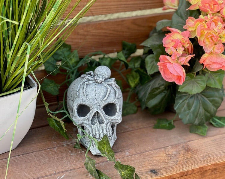 Totenkopf-Gartenfigur mit einer Spinne auf dem Kopf, dekoriert auf Holz mit Efeublättern und Topfpflanzen.