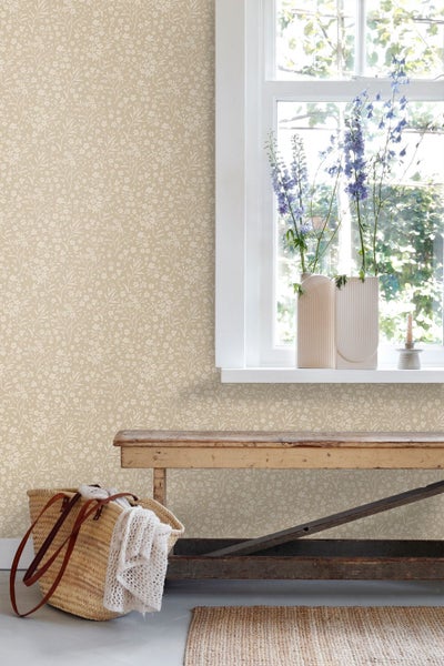 Wand mit beiger Blumentapete, einer rustikalen Holzbank, einer Strohtasche und einem Fenster mit Blumen in strukturierten Vasen.