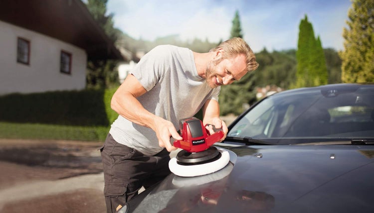 Ein Mann poliert die metallene Motorhaube eines Autos mit einer Einhell Autopoliermaschine in einer hellen Außenumgebung.