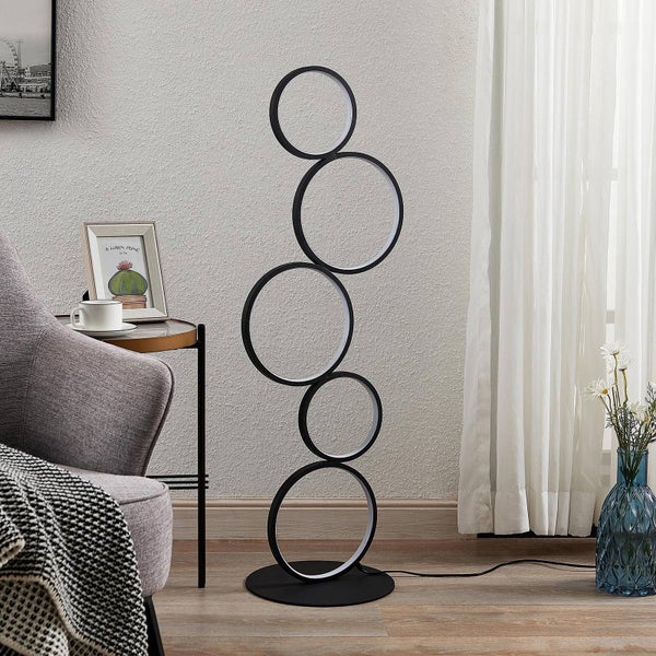 Moderne LED-Stehleuchte mit fünf ineinandergreifenden Ringen in einem hellen Wohnzimmer neben einem grauen Sessel.