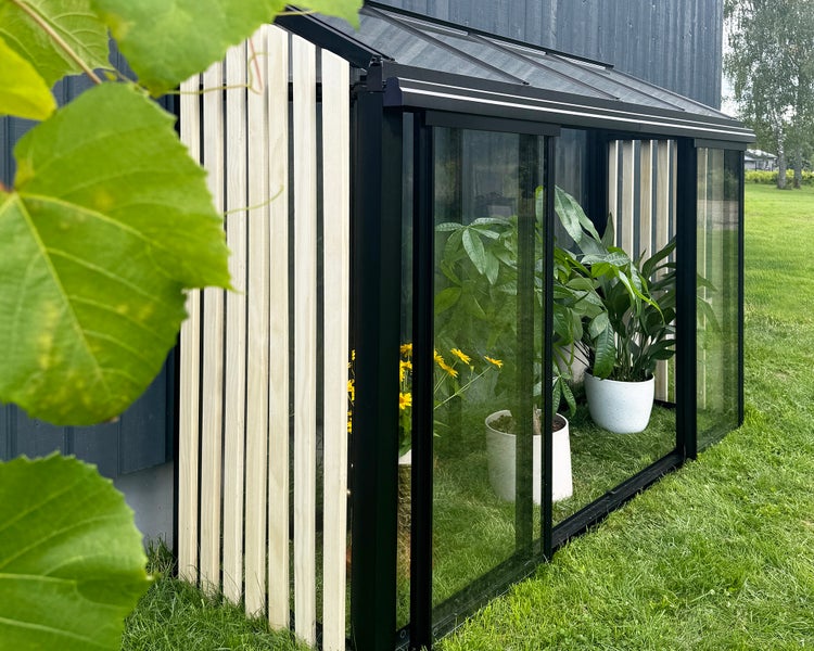 Anlehngewächshaus mit schwarzem Aluminiumrahmen, Glasschiebetüren und Seitenwand aus Holzlamellen, gefüllt mit Topfpflanzen auf einer Rasenfläche.
