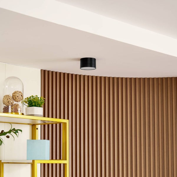 Schwarze zylindrische Deckenleuchte montiert an der Decke über einer Wand mit Holzlamellen und einem goldenen Regal.