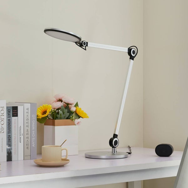 Moderne silberne LED-Schreibtischleuchte mit verstellbarem Arm und rundem Kopf auf einem hellen Schreibtisch mit Dekoration.