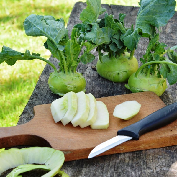 Frischer Kohlrabi mit grünen Blättern auf einem rustikalen Holztisch, teilweise aufgeschnitten auf einem Holzschneidebrett neben einem Messer.