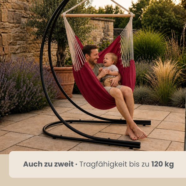 Ein Vater und sein Kleinkind sitzen gemeinsam in einem Hängesessel mit Gestell auf einer Steinterrasse im Garten.