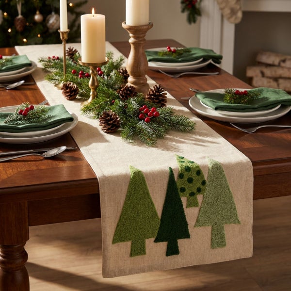 Festlich gedeckter Holztisch mit Tischläufer mit Tannenbaummotiven, Tannengrün, Zapfen und Kerzen für eine weihnachtliche Tischdekoration.
