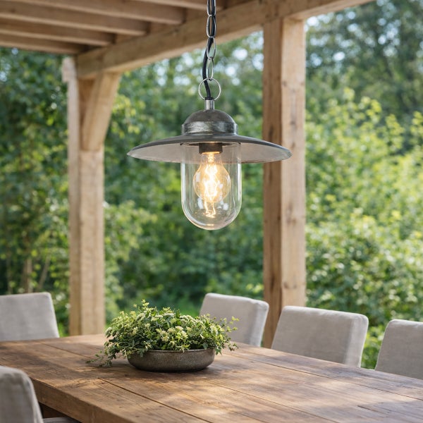 Rustikale Außenhängeleuchte aus Metall und Glas über einem Holztisch auf einer überdachten Terrasse mit Blick ins Grüne.