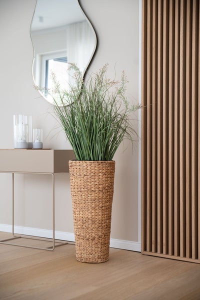 Großer geflochtener Pflanzkübel mit künstlichem Gras in einem hellen, modernen Zimmer mit Spiegel, Konsolentisch und Holzpaneelen.