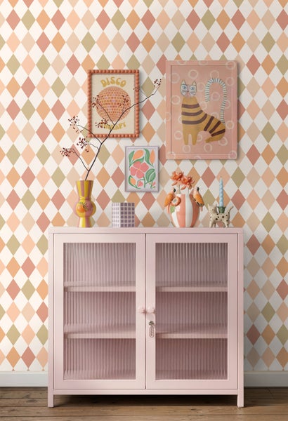 Vliestapete mit Harlekinmuster in Orange und Beige hinter einer rosa Vitrine mit geriffeltem Glas, dekoriert mit Vasen und gerahmten Wandbildern.