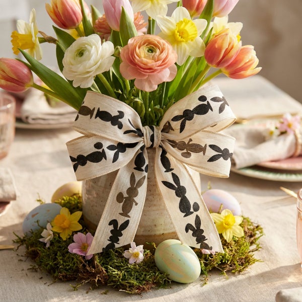 Ostergesteck mit Tulpen und Narzissen in einer Vase, verziert mit einer Schleife mit Hasenmuster und umgeben von Moos sowie Ostereiern.