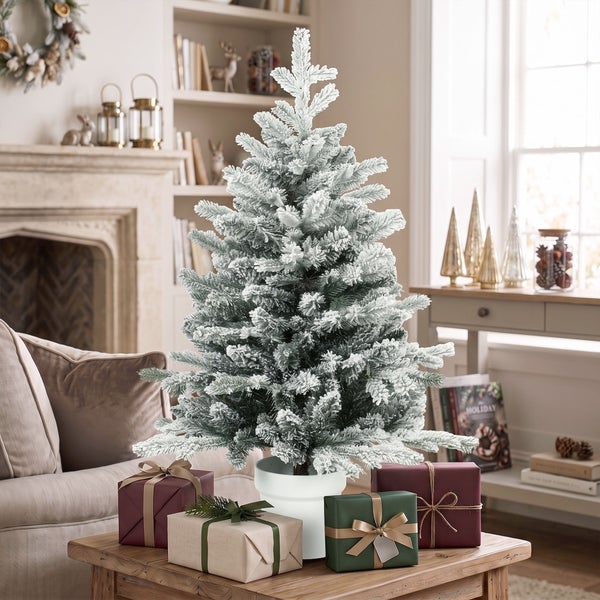 Künstlicher, beschneiter Weihnachtsbaum im Topf auf einem Holztisch, umgeben von Geschenkpaketen in einem gemütlichen Wohnzimmer.