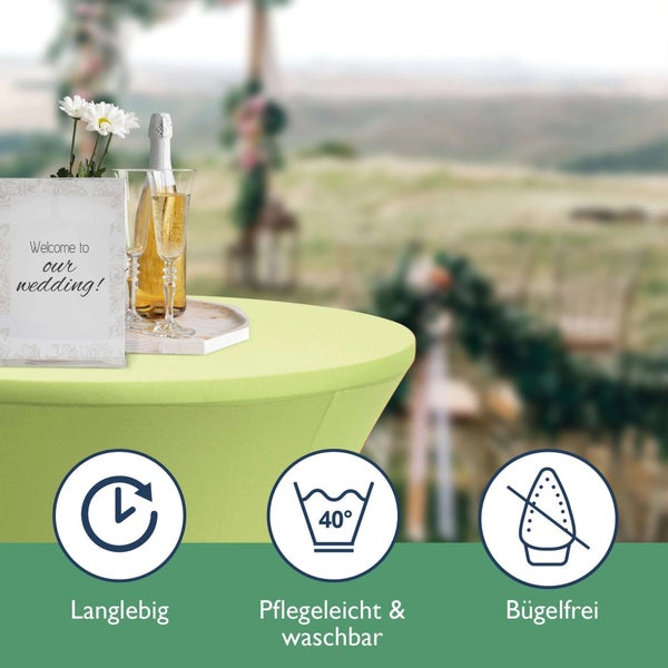 Szene mit einem Tisch mit grüner Stretch-Tischdecke, einem Schild mit der Aufschrift Willkommen zu unserer Hochzeit, Champagner und Symbolen für Langlebigkeit, Pflegeleicht und Waschbarkeit sowie Bügelfreiheit.