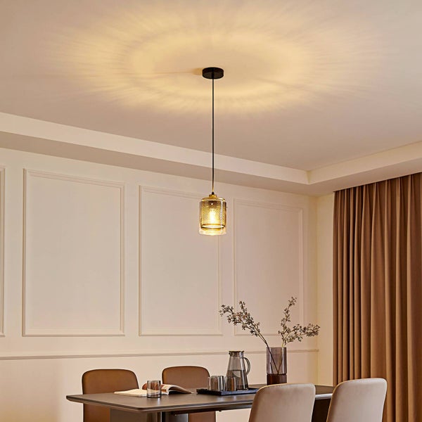 Moderne Pendelleuchte mit Metallgitter und Glaszylinder über einem Esstisch in einem hellen Raum mit dekorativen Wandpaneelen.