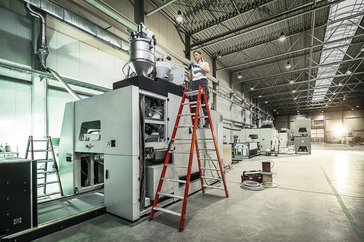 Eine Arbeiterin steht auf einer Stehleiter und führt Wartungsarbeiten an einer großen Industriemaschine in einer Lagerhalle durch.