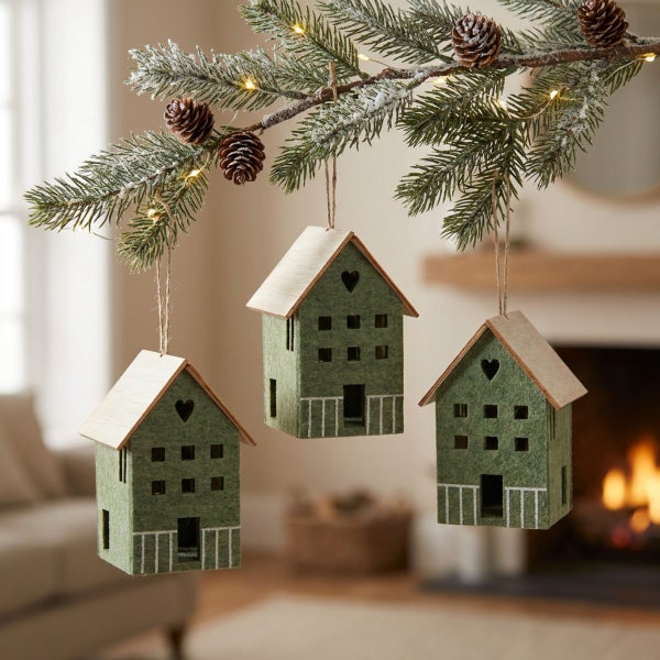 Drei kleine, hängende Weihnachtsanhänger in Form von Häusern aus Filz und Holz, befestigt an einem weihnachtlich geschmückten Tannenzweig.