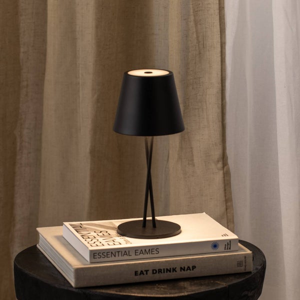 Schwarze Akku-Tischleuchte mit kegelförmigem Schirm auf Büchern vor Vorhängen in einem wohnlichen Raum.