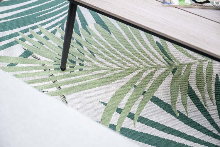 Nahaufnahme eines Outdoor-Teppichs mit Palmenblatt-Muster unter einem hölzernen Gartentisch.
