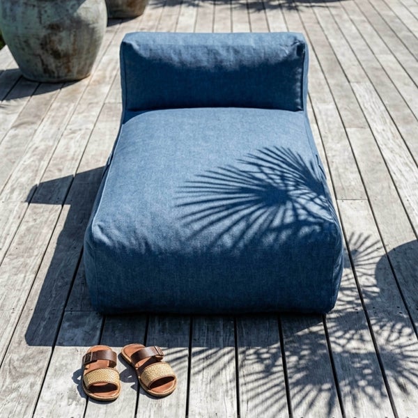 Blaue gepolsterte Gartenliege aus Stoff auf einer Holzterrasse mit Palmenschatten, Sandalen und Pflanzgefäß in heller Atmosphäre.