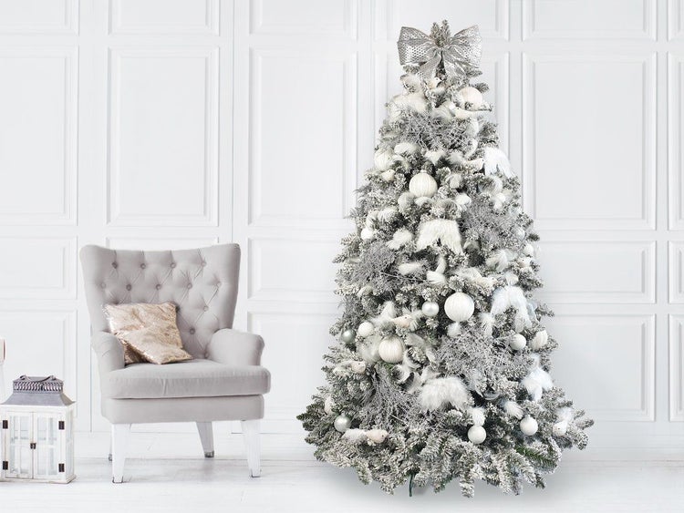 Weihnachtsbaum mit Kunstschnee dekoriert mit Kugeln und Schleife in einem hellen Wohnraum mit Polstersessel und Laterne.