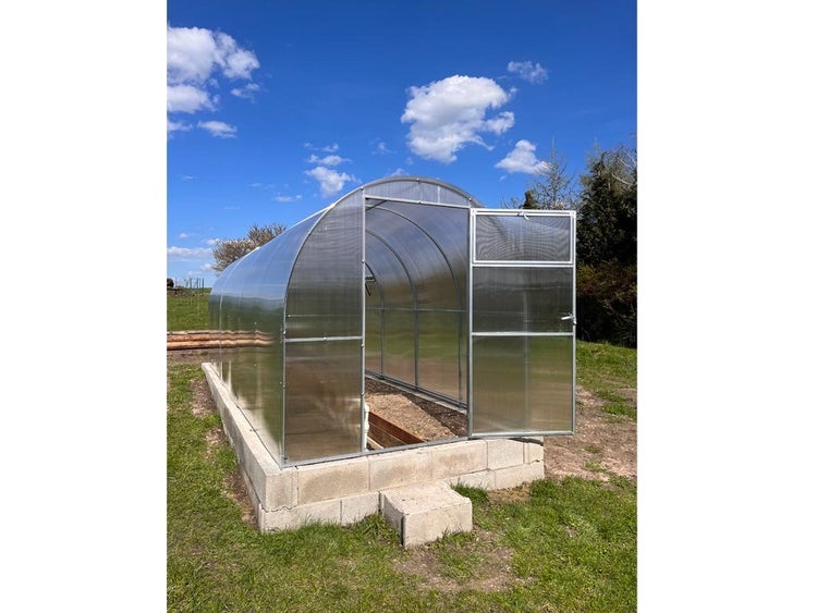 Gewächshaus aus Polycarbonat mit geöffneter Tür auf Betonfundament im Garten.